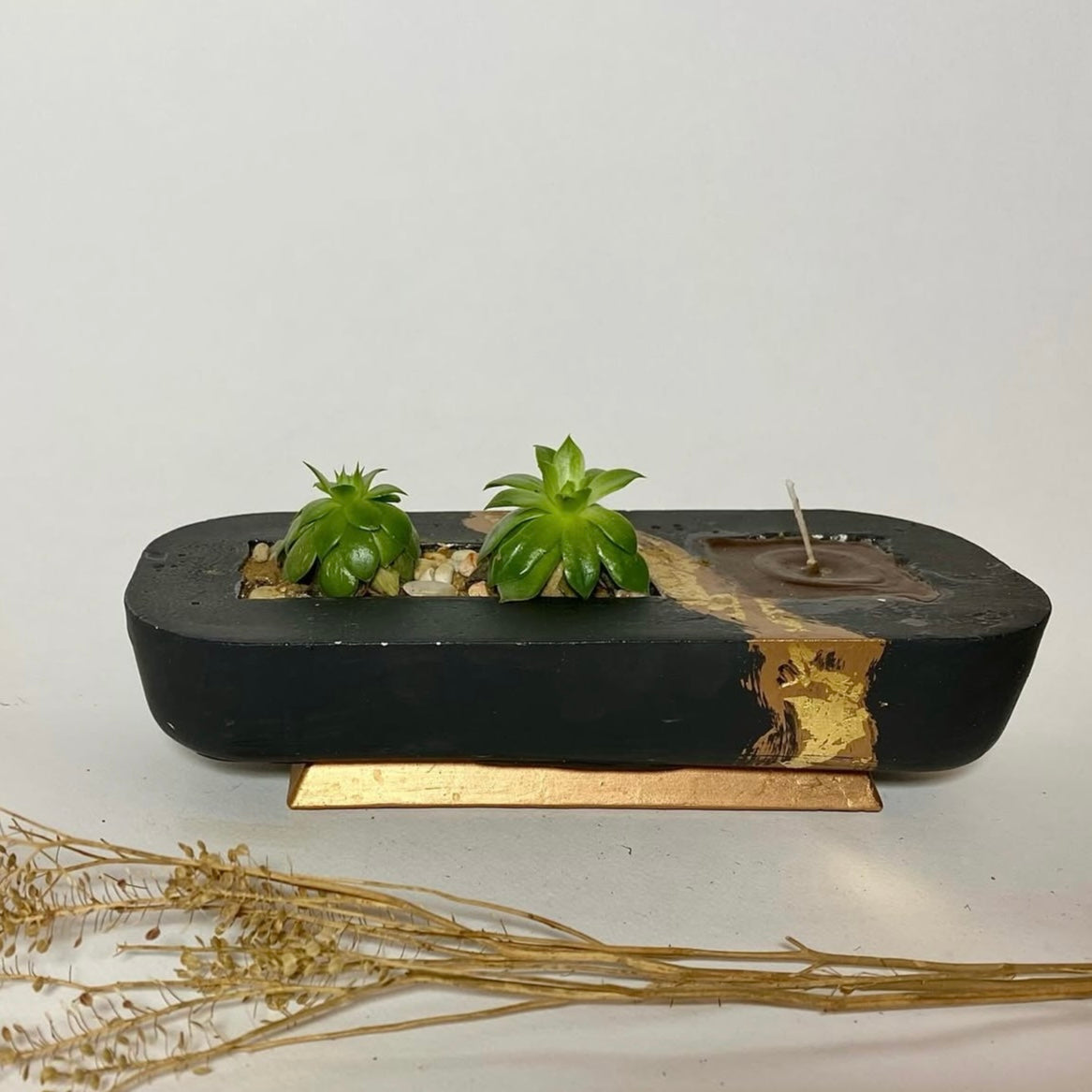 A rectangular black plant pot with a section containing a small green plant and decorative stones. There is also a small brown structure resembling a candle holder.