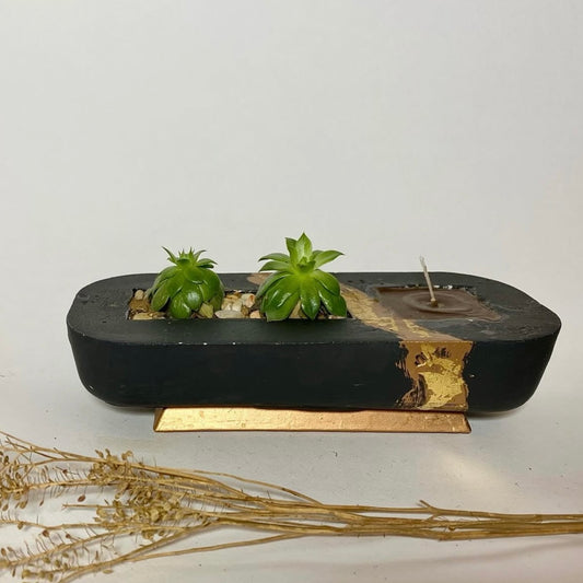 A rectangular black plant pot with a section containing a small green plant and decorative stones. There is also a small brown structure resembling a candle holder.