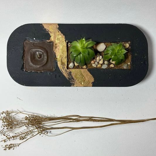 A rectangular black plant pot with a section containing a small green plant and decorative stones. There is also a small brown structure resembling a candle holder.